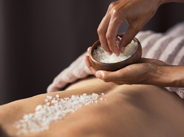 Masseuse hands doing massage on woman's back at beauty salon. Beauty therapist pouring salt scrub on woman back at health spa. Scrubbing and skin care concept.