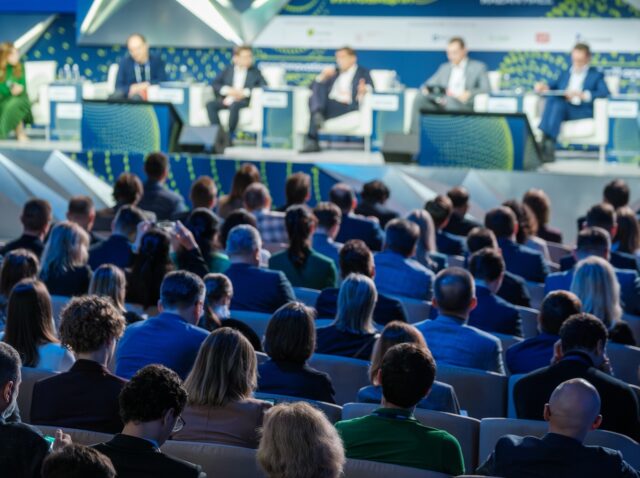 Large business conference with a panel of speakers and a diverse audience in attendance, focusing on various topics and discussions.